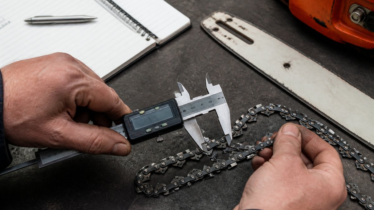 Hands using digital calipers to measure chainsaw part stock pitch and gauge on a workbench.
